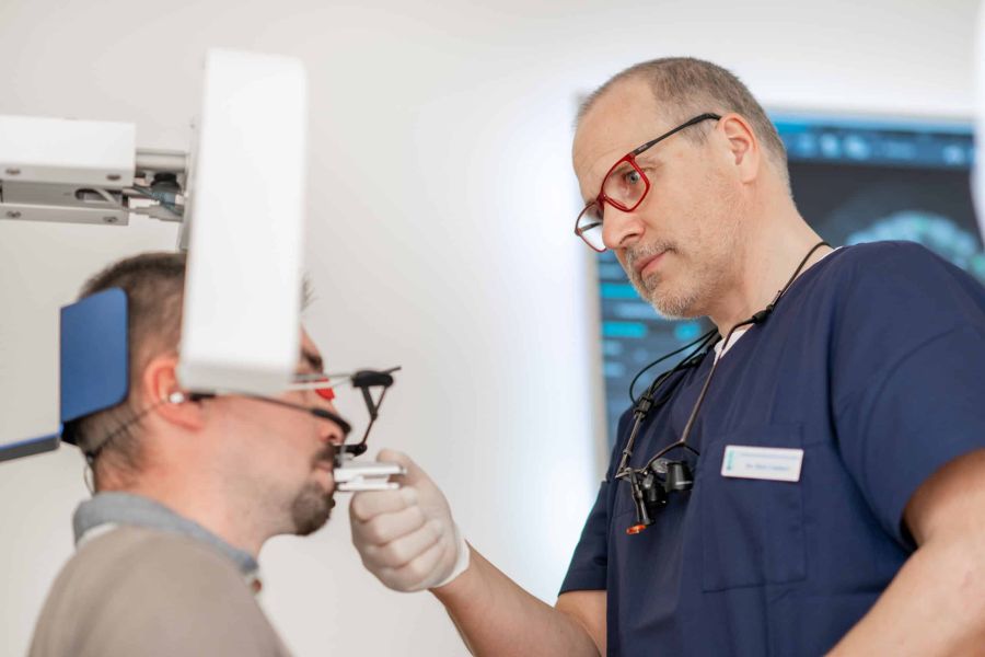 Zahnarzt in blauer Berufskleidung passt einem Patienten einen Gesichtsbogen zur zahnmedizinischen Vermessung an.