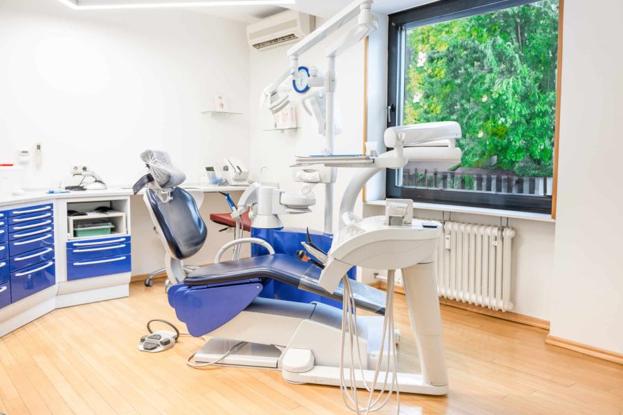 Modernes Behandlungszimmer in unserer Zahnarztpraxis mit blauem Behandlungsstuhl, zahnmedizinischen Geräten und großem Fenster mit Blick ins Grüne.