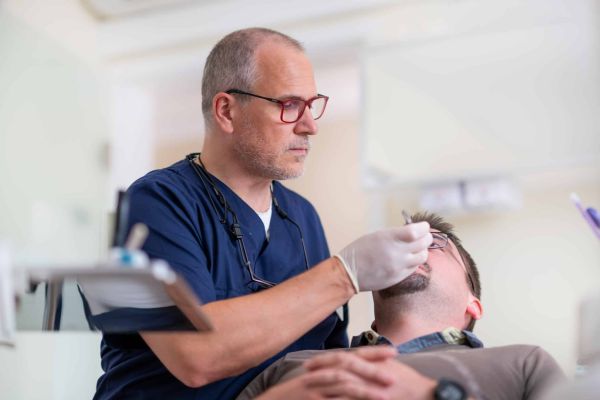 Zahnarzt in blauer Berufskleidung untersucht einen liegenden Patienten mit einem zahnmedizinischen Instrument.