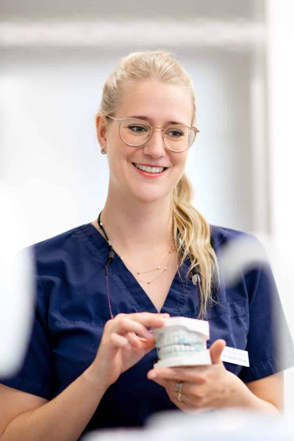 Frau Dr. Völlings in blauer Praxisbekleidung hält ein Modellgebiss und lächelt eine Patientin an.