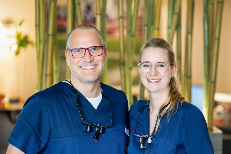 Dr. Dirk Lindner und Dr. Antonia Völlings in blauer Praxisbekleidung mit Lupenbrillen, lächelnd in die Kamera.