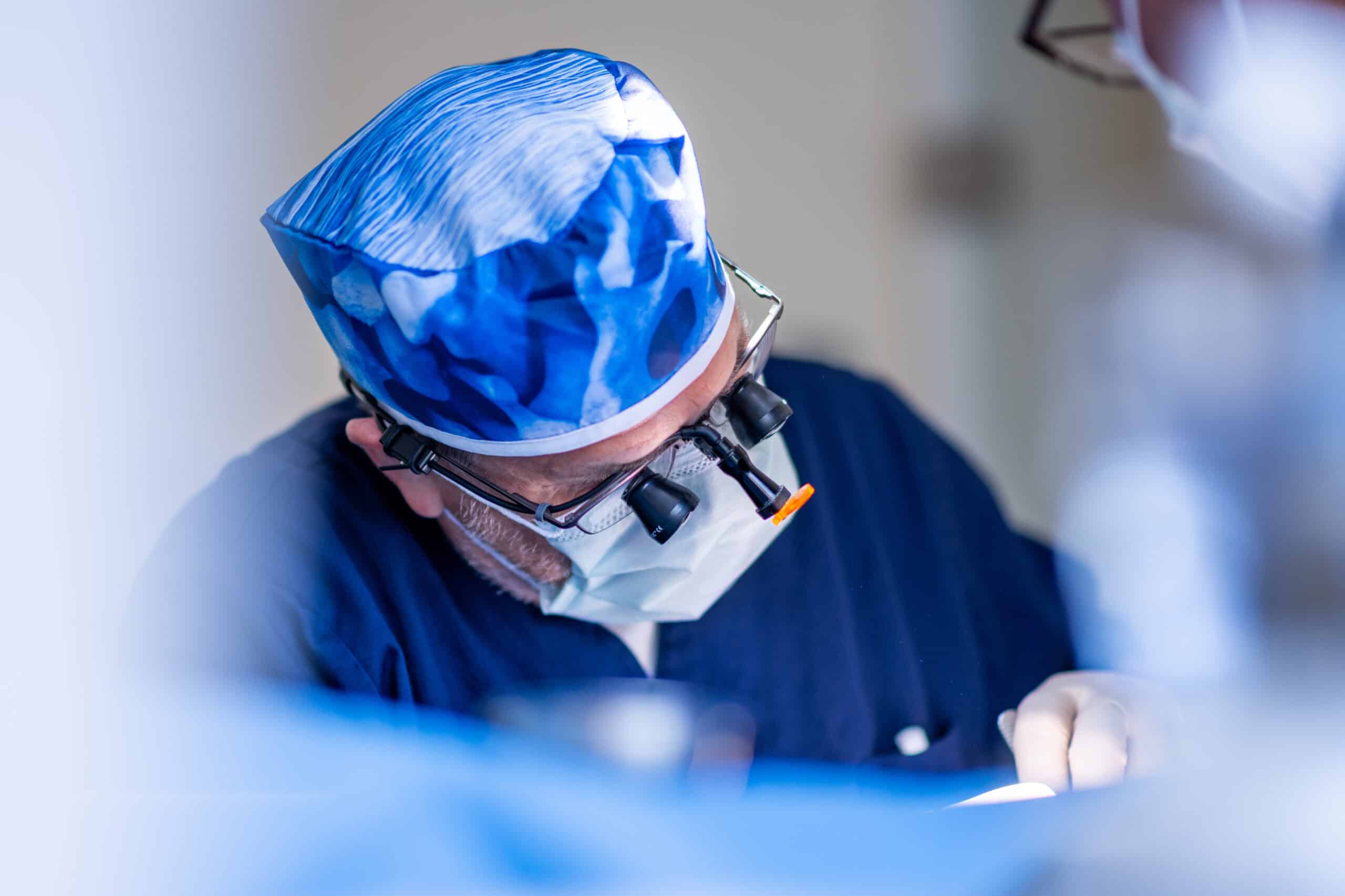 Zahnarzt mit Lupenbrille, OP-Haube und Maske führt konzentriert einen chirurgischen Eingriff durch. Sterile Umgebung und professionelle Ausstattung.